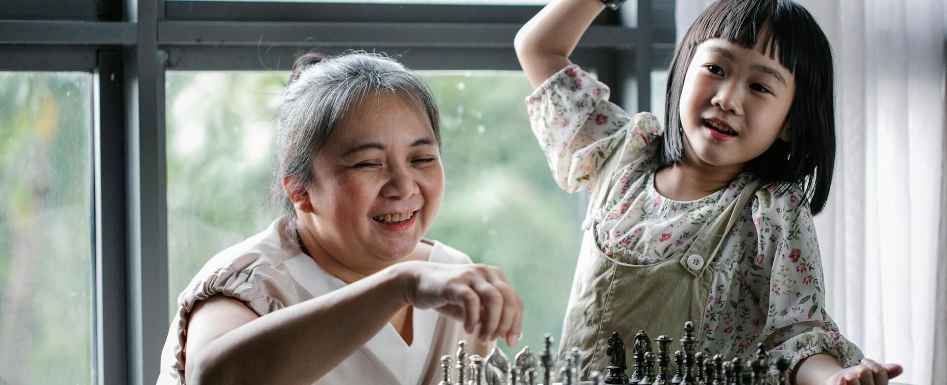 cheerful senior ethnic woman with cute granddaughter playing chess at home