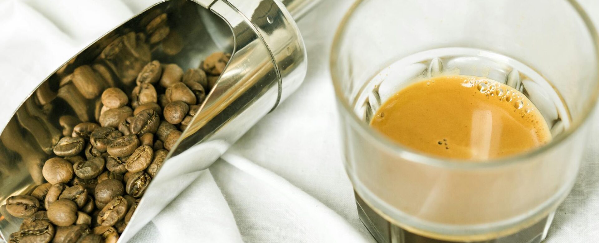 a clear drinking glass with coffee near the stainless scoop with coffee beans